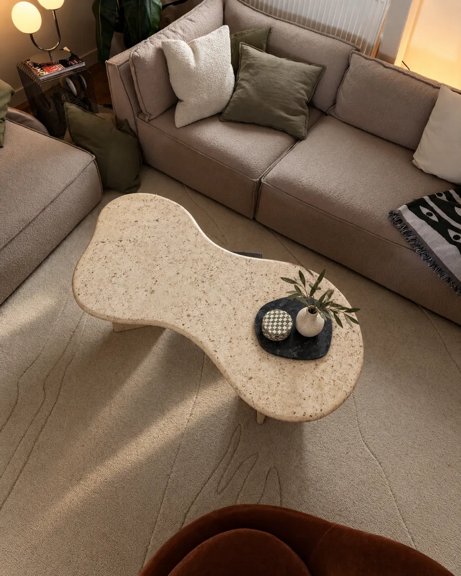 The stone coffee table from above, an organic shape in light beige quartzite on a rug in front of a sofa
